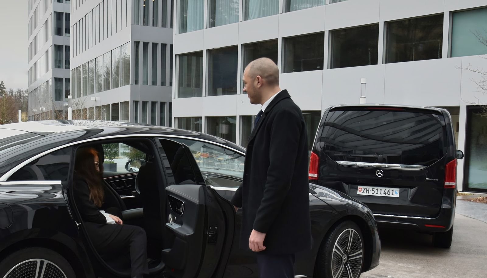 Chauffeur hält einem Gast die hintere Tür eines schwarzen Mercedes EQE auf, dahinter eine Mercedes V-Klasse
