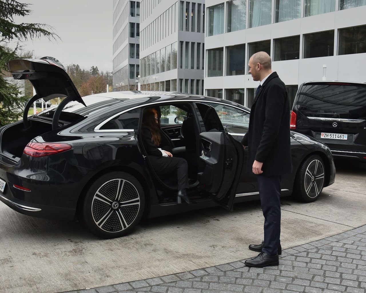 Chauffeur holding the rear door of a black Mercedes EQE, a V-Class behind