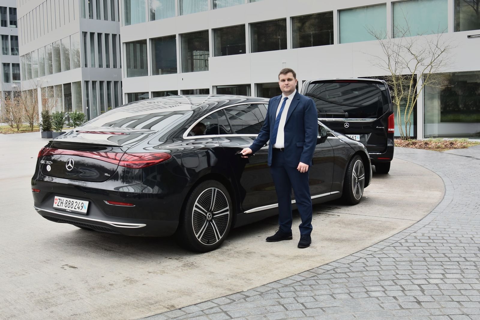 Express Transfer chauffeur in a suit beside a black Mercedes EQE, with a Mercedes V-Class behind