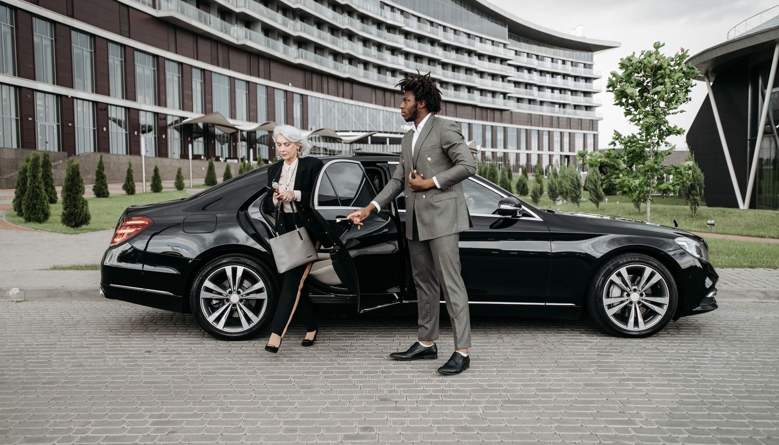 Chauffeur in a suit holding the rear door of a black sedan for a guest outside a modern hotel