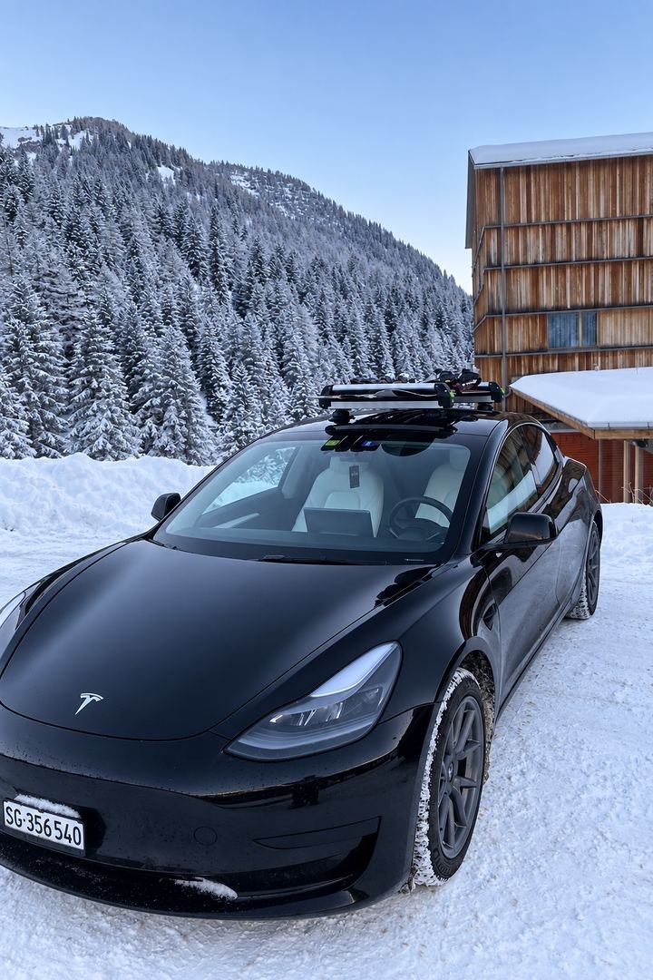 Our black Tesla Model 3 with a roof ski carrier, parked in the snow below a forested mountain