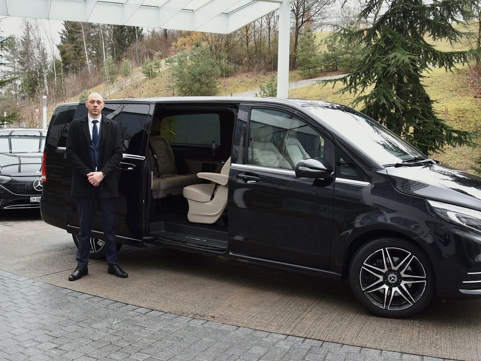 Chauffeur im dunklen Anzug neben einer schwarzen Mercedes V-Klasse mit geöffneter Schiebetür